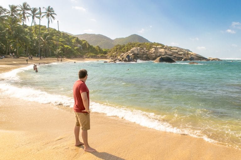 Jeremy Storm in a red shirt standing on a beach in Tayrona National Park Colombia--be sure to go through this international travel checklist before heading off to gorgeous spots like this!