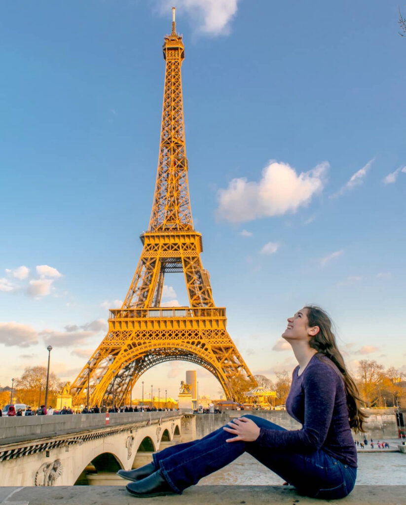 kate storm sitting on a wall along the seine in paris looking up at the eiffel tower during her second time in paris france