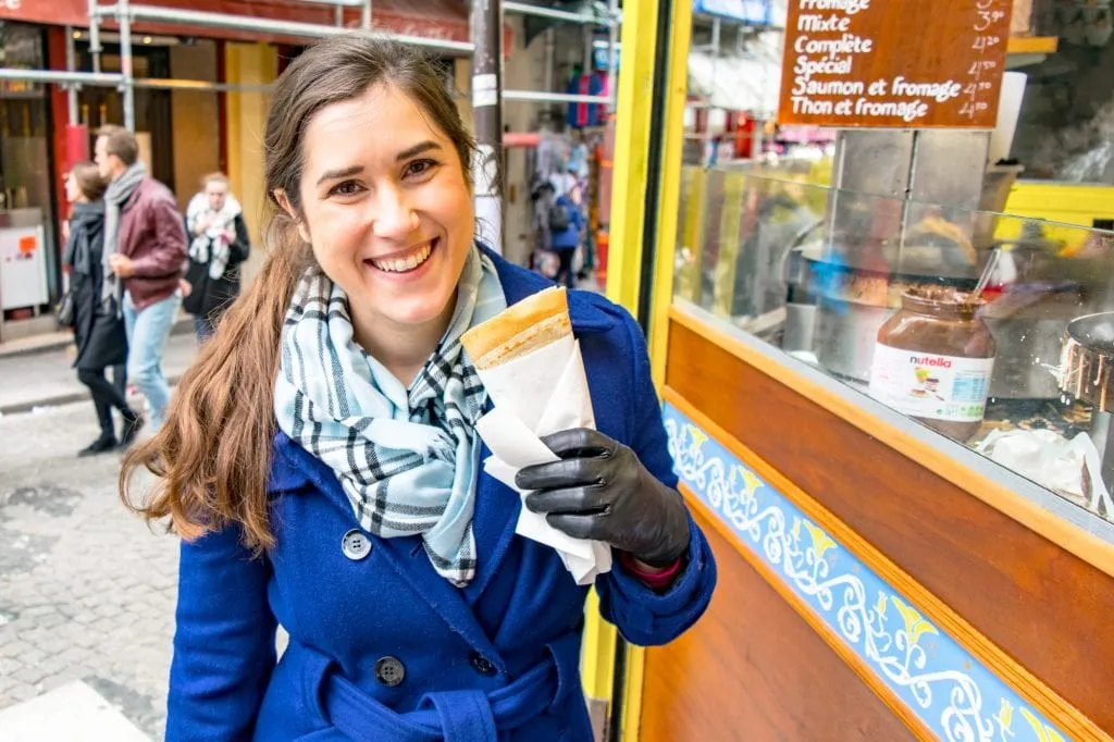 Kate Storm in a blue coat holding a crepe while sightseeing in Paris--any weekend trip to Paris definitely requires eating at least a couple of crepes!