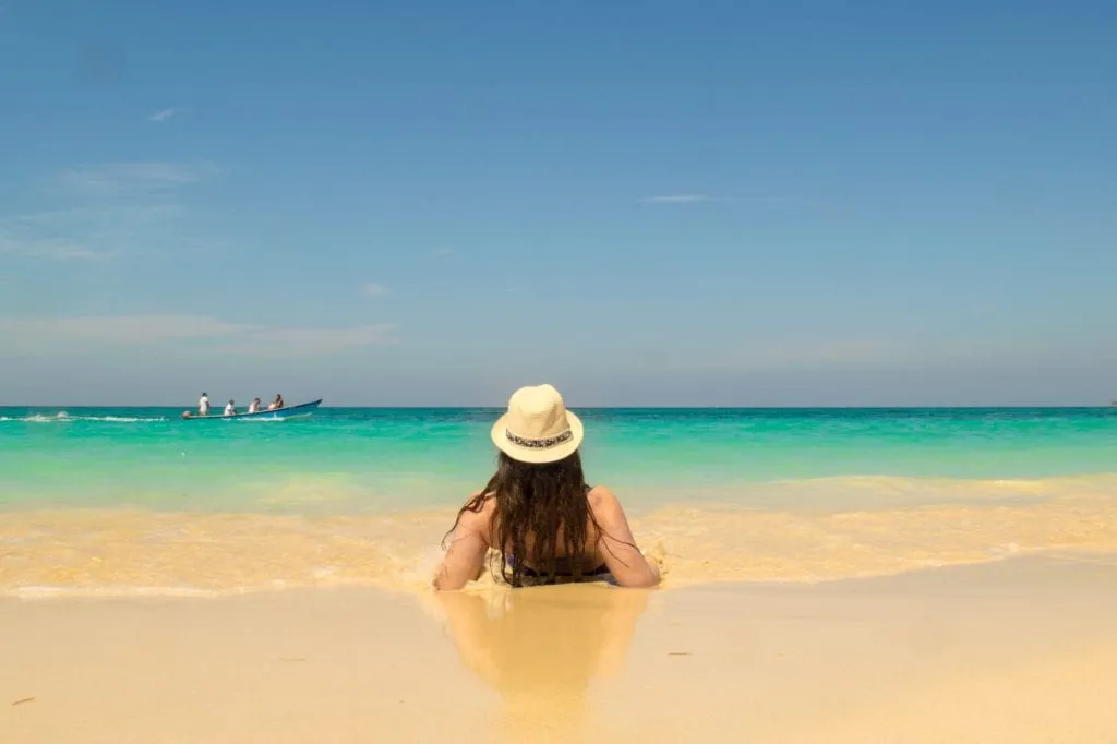 Kate Storm in a sun hat laying down on Playa Blanca Beach near Cartagena and looking out toward the water. There's a boat in the distance.