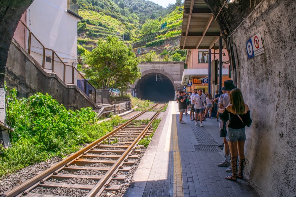 Photo of the empty train tracks at a station in Cinque Terre. Some people are standing to the side and waiting on the platform.