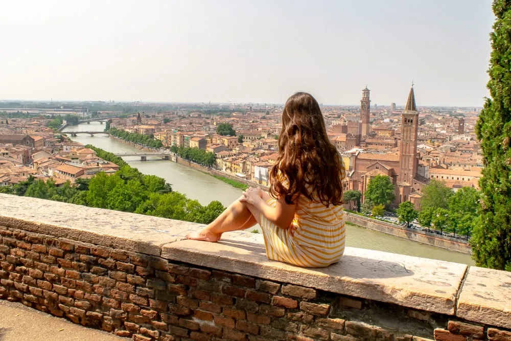 Kate in a yellow dress sitting on a wall at Castel San Pietro overlooking the Adige River and skyline of Verona. Verona is an excellent place to visit on your Italy honeymoon!