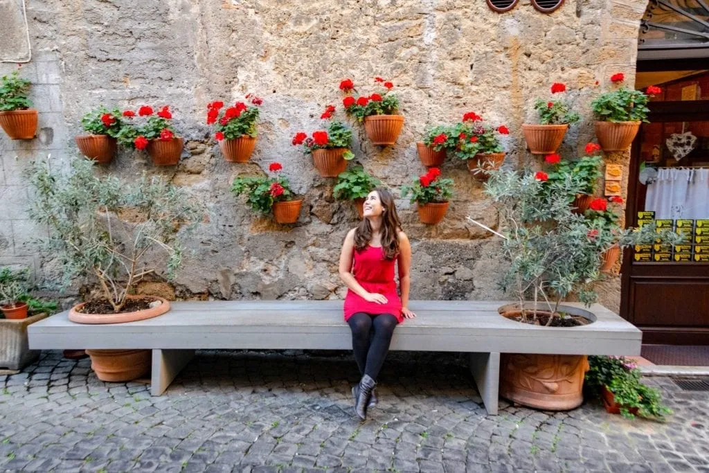 Kate sitting on a bench in Orvieto Italy. She's wearing a red dress, black tights, and black boots. There are red flowers in pots on the wall behind her.