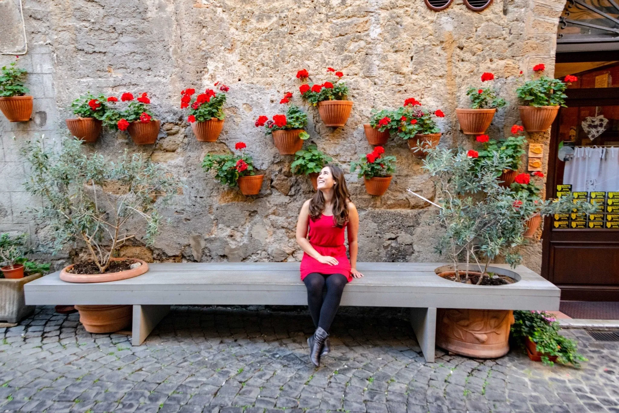 Kate Storm sitting on a bench in Orvieto Italy. She's wearing a red dress, black tights, and black boots. There are red flowers in pots on the wall behind her.