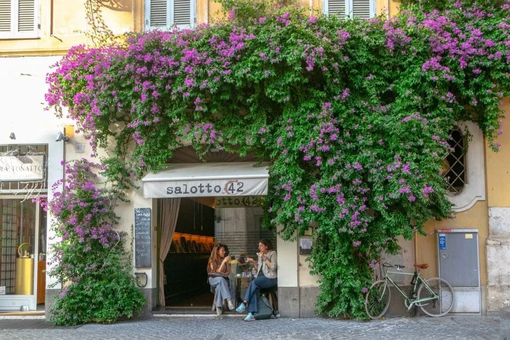 two women sitting at a cafe in rome italy with purple flowers blooming above them