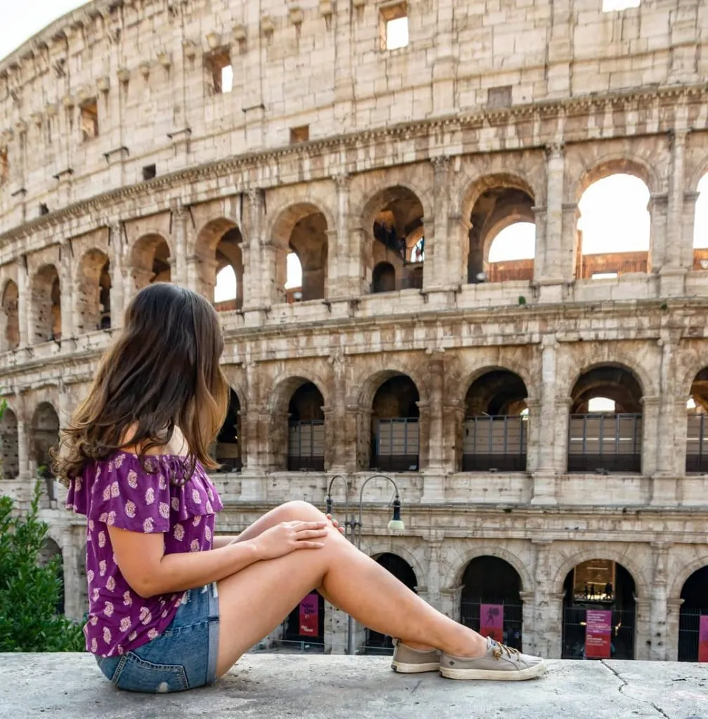 kate storm in a purple shirt sitting in front of the colosseum, one of the top attractions on an itinerary for rome italy