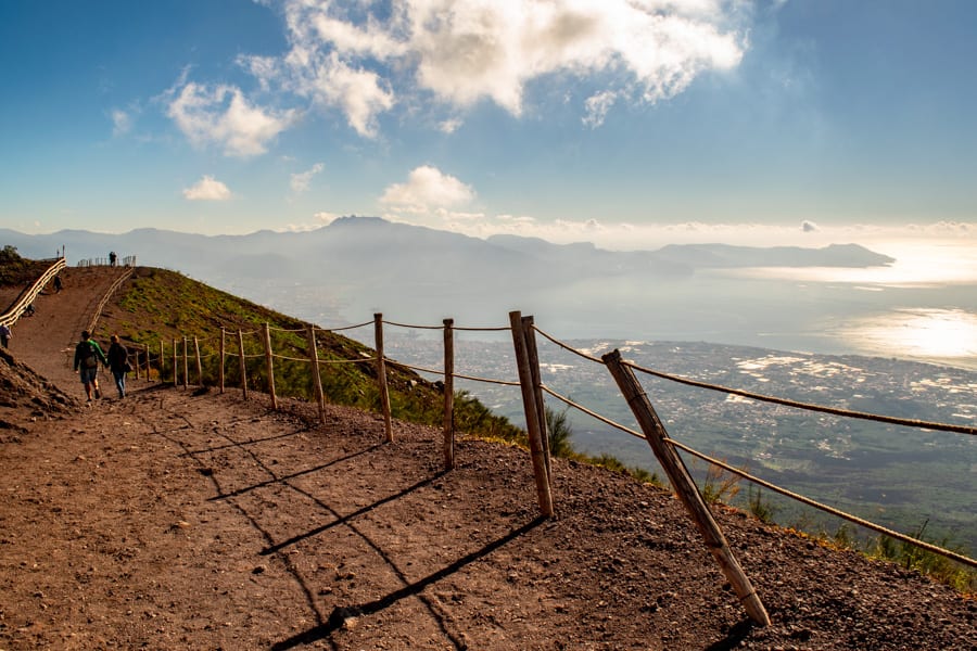 Photo of trail along the crater of Vesuvius, with distant view of Capri on the upper right. There's a crooked wooden hand rail.