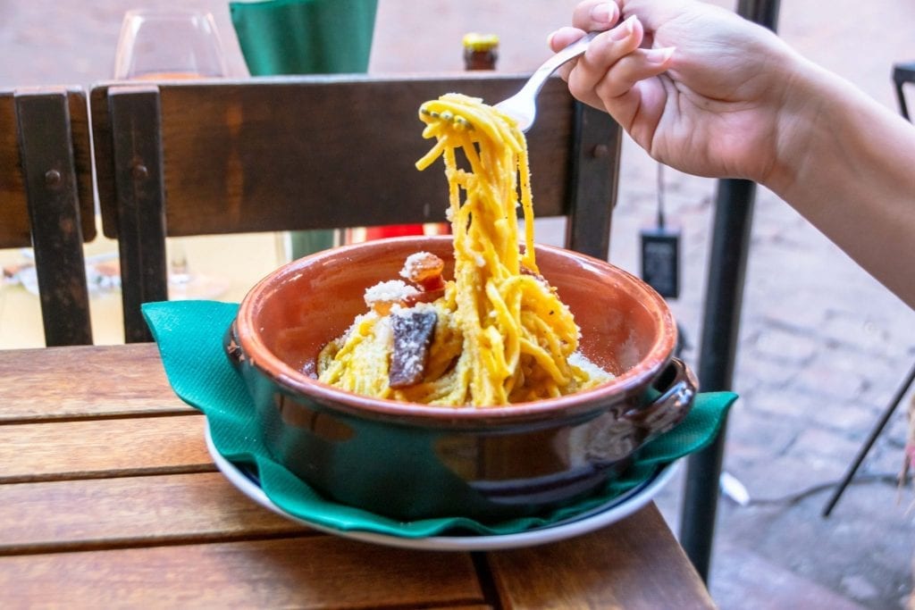 Photo of a bowl of carbonara, a woman's hand is holding up one bite with a fork. Eating plenty of this is definitely among my personal favorite travel tips for Italy.