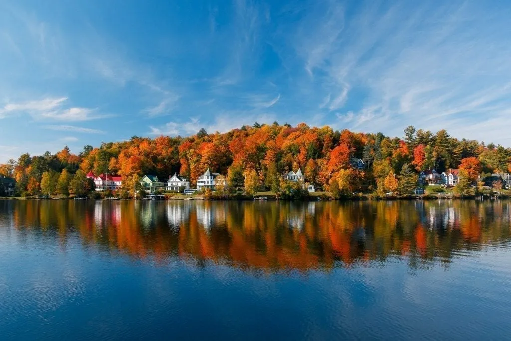 Saranac Lake, Adriondacks New York in the fall, with houses nestled against the lake shore, taken from across the water. Upstate NY in the fall in is one of the most romantic USA trips to take!