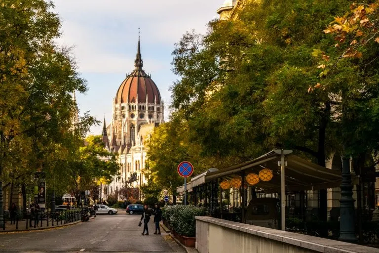 Photo of a street in Budapest lined by leafy green trees. Dome of Hungarian Parliament is in the distance.