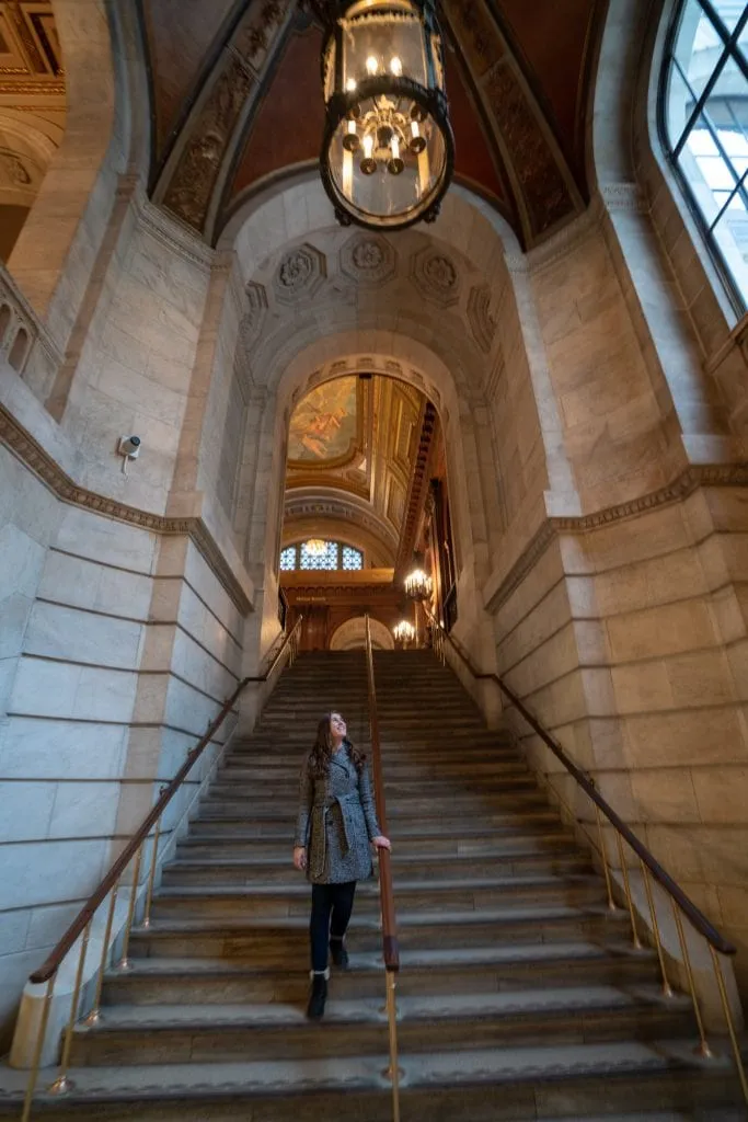 Kate walking down a stone staircase in the NYC Public Library, 5th Avenue branch. Kate is wearing a gray coat.