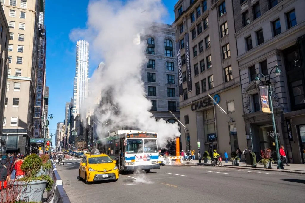view of a smoking grate on a street in new york city with a taxi and bus driving by