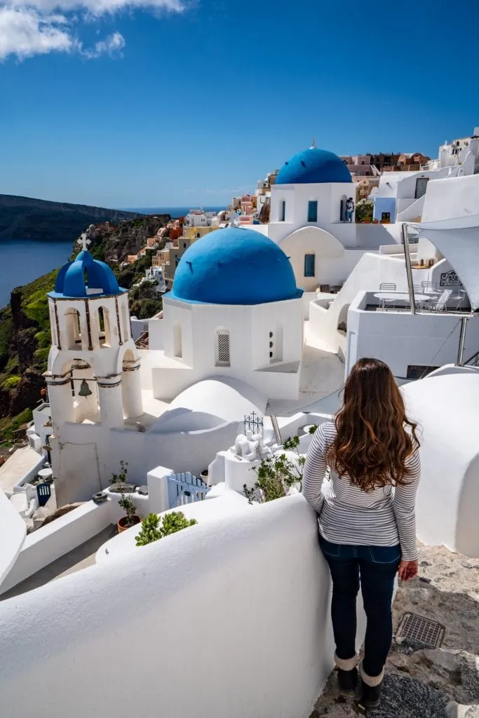 Kate Storm in a white shirt and blue jeans overlooking the blue domes of Oia in Santorini--this island is incredible and worth visiting as part of your 2 weeks in Europe!