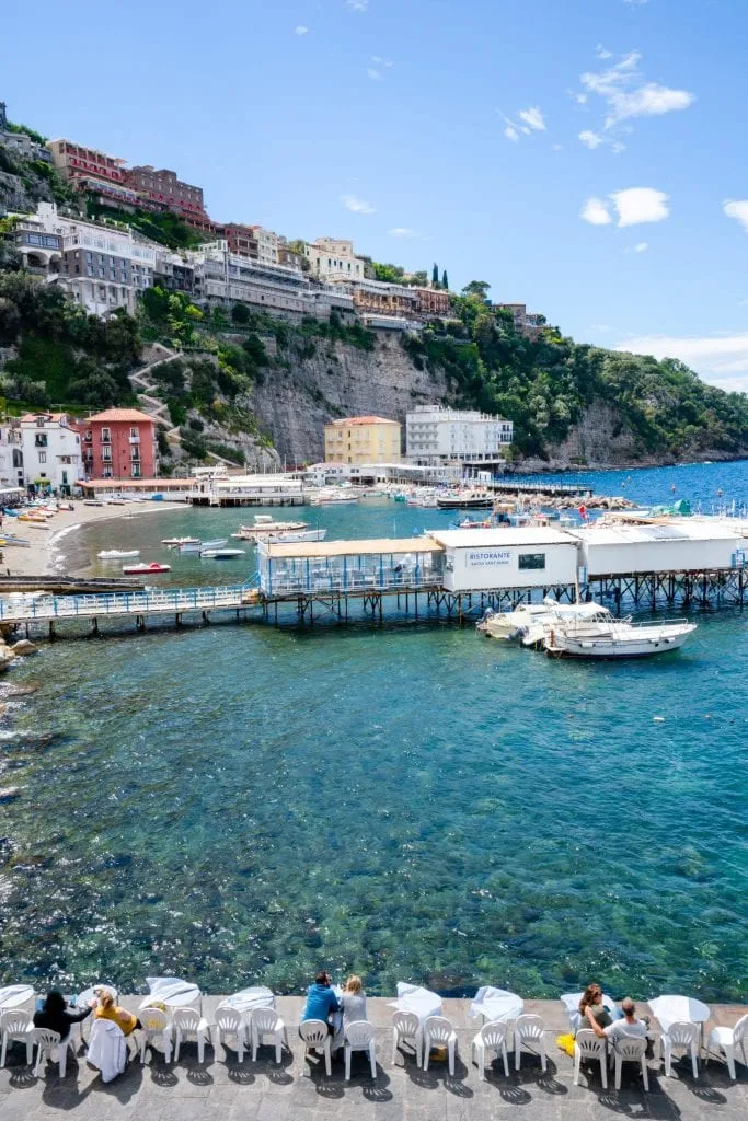 View of Marina Piccola, Sorrento, with white tables with people eating at them in the foreground. If you have extra time for your Amalfi Coast itinerary, be sure to see Sorrento.