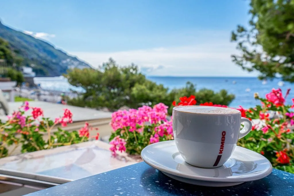 View of pink and red flowers with Positano beach in the background and cup of coffee on a white saucer in the foreground.