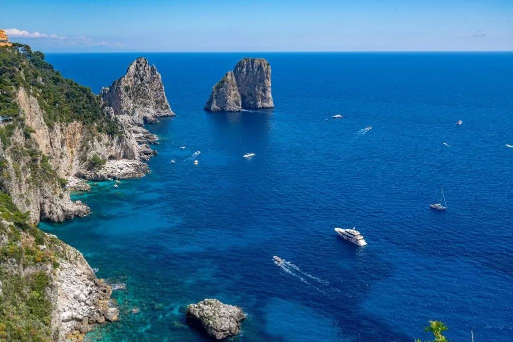 Photo of the Faraglioni of Capri taken from the Garden of Augustus. There are boats in front of the Faraglioni and the cliffs of Capri to the left.