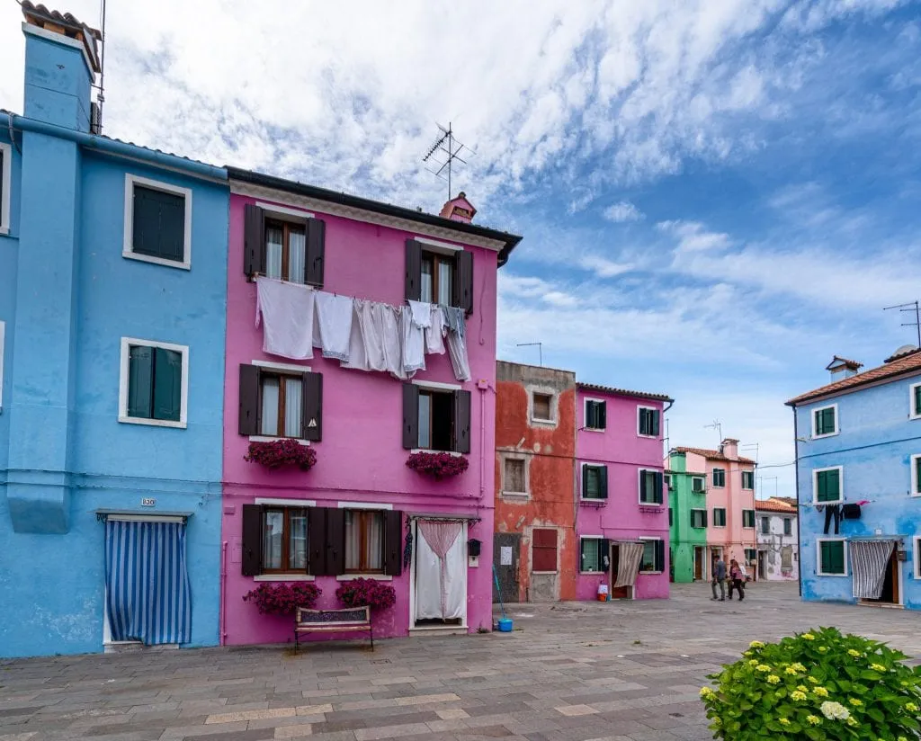 Photo of colorful houses in Burano as seen from a piazza. Laundry is hanging from some windows.