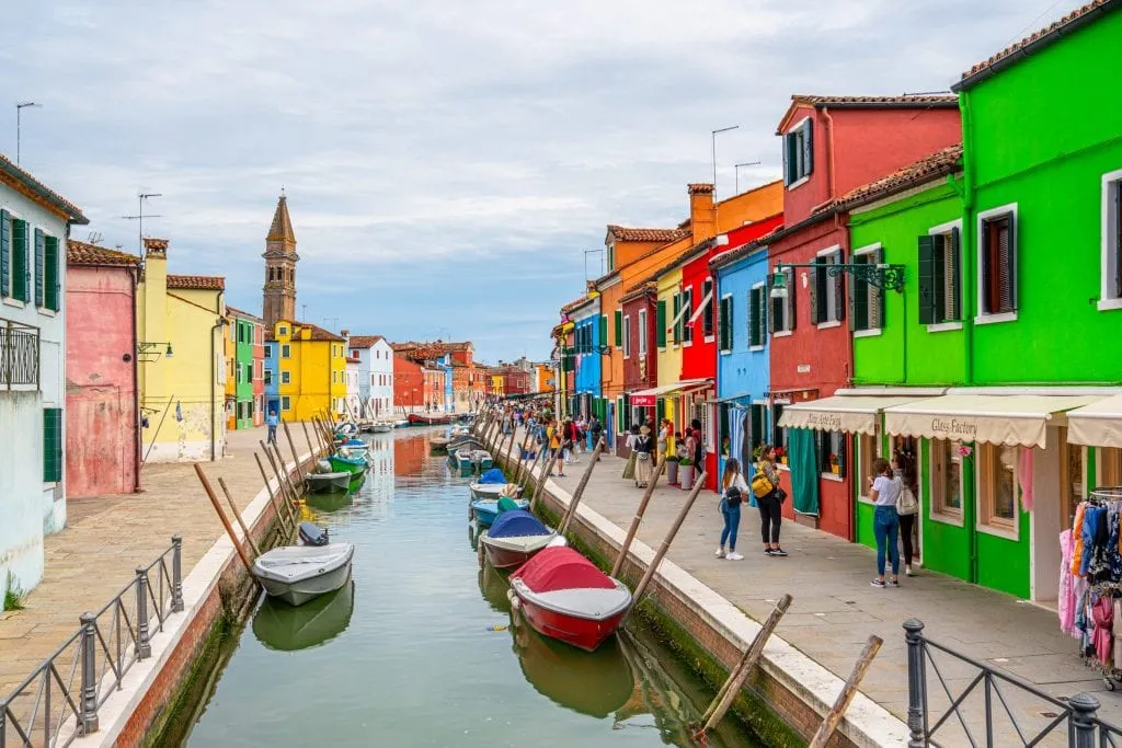 Photo of a canal in Burano with colorful houses on both sides and leaning tower in the background.