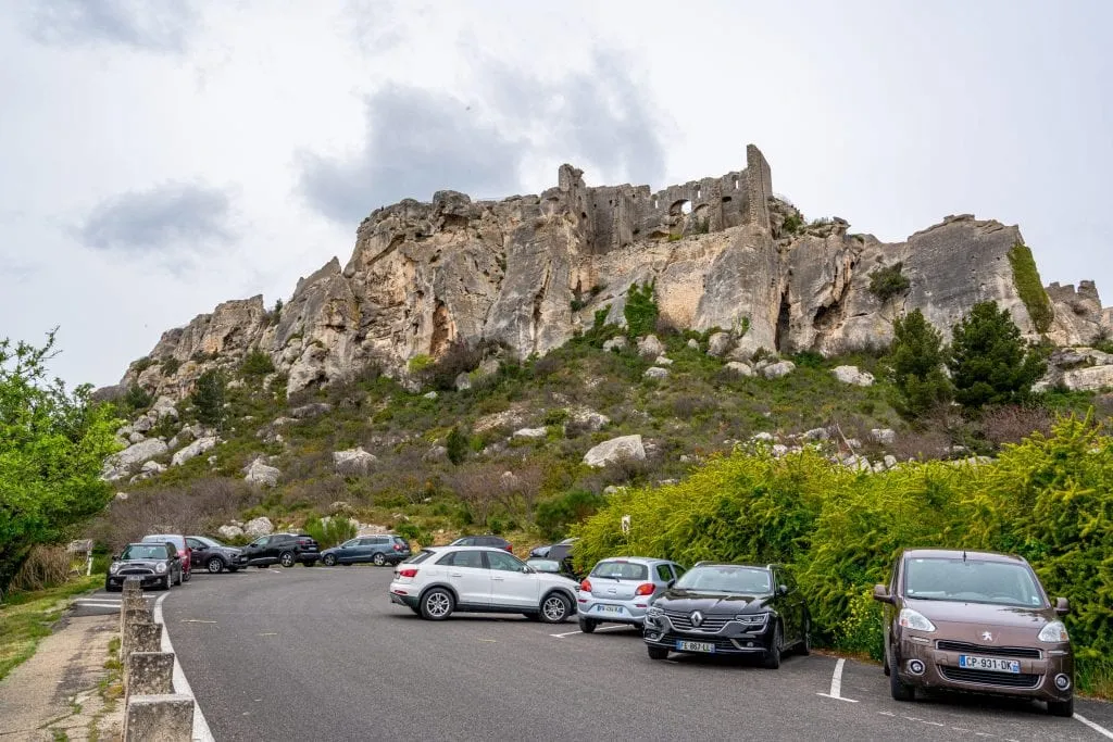 Parking lot outside of Les Baux-de-Provence. Several cars are parked to the right and the city is visible in the background.