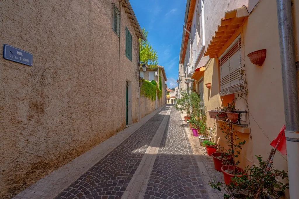 Empty pedestrian street in Cassis France. There's a blue street sign on the left side of the photo and red flowers on the right.