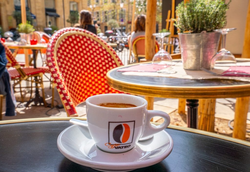 Cup of coffee sitting on an outdoor table at a cafe in Aix-en-Provence. There's a red and white checkered chair behind the coffee.