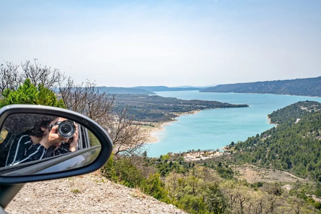 Photo of Lake Sainte-Croix with Kate visible snapping a photo in the rearview mirror on the left--a camera for photos like this definitely belongs on your road trip packing list!