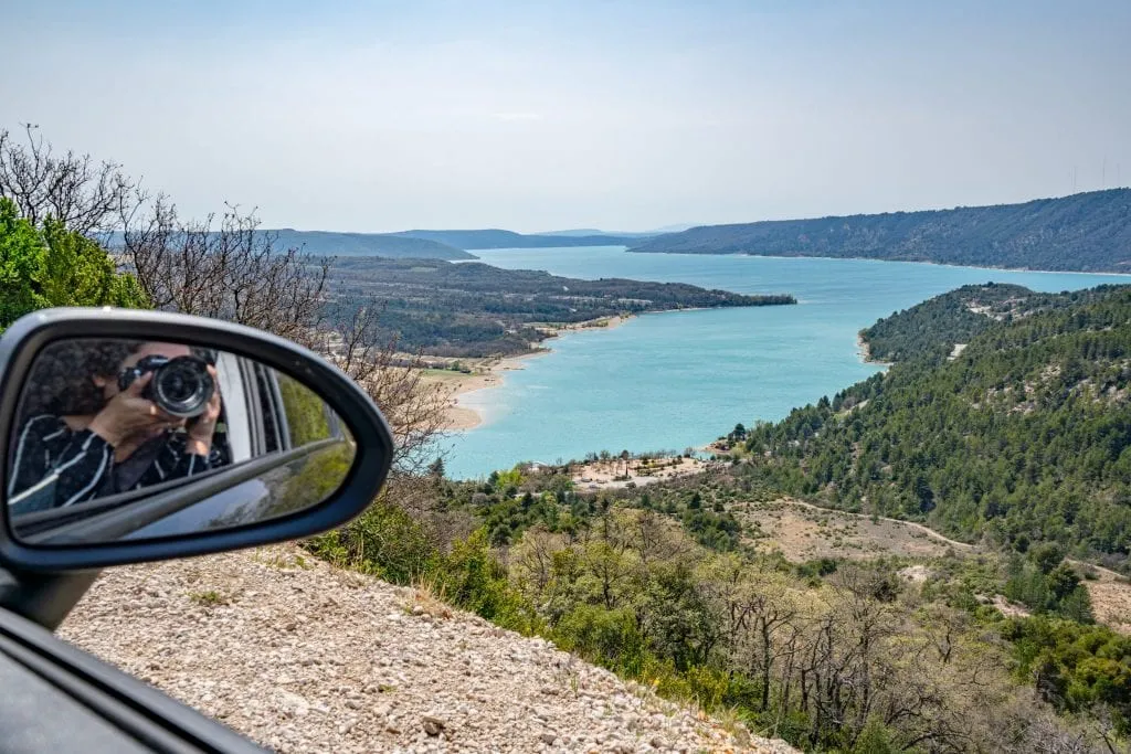 Photo of Lake Sainte-Croix as seen while driving in France. You can see Kate holding a camera to her face reflected in the rearview mirror of our rental car on the left side of the photo