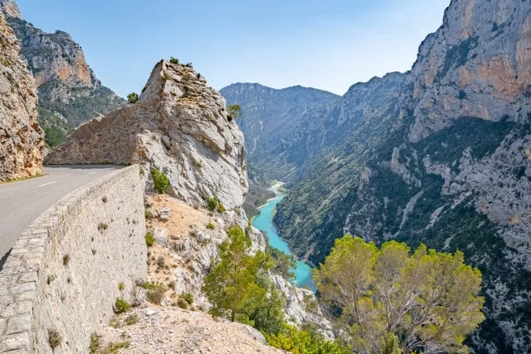 Photo of an empty road on the left, with the Verdon Gorge to the right. The turquoise river of the gorge is visible in the center of the photo. Definitely worth stopping here on a France road trip!