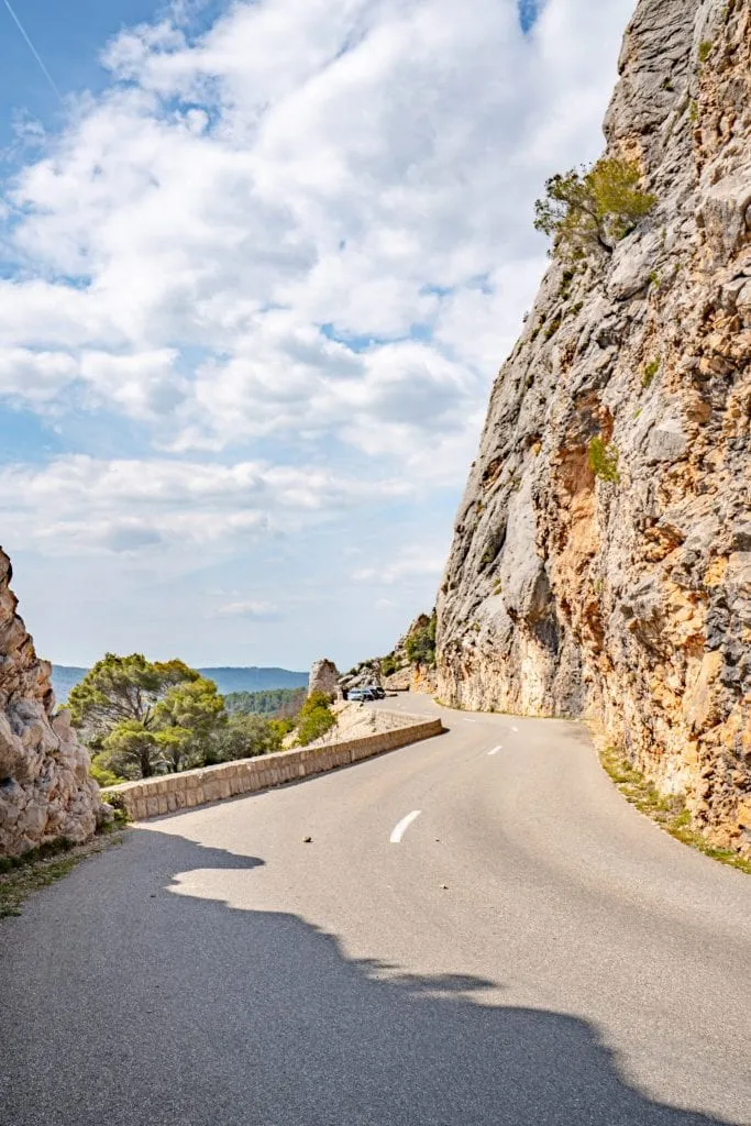 Vertical photo of a winding road in France. There's a dotted white line in the center and a large cliff to the right.