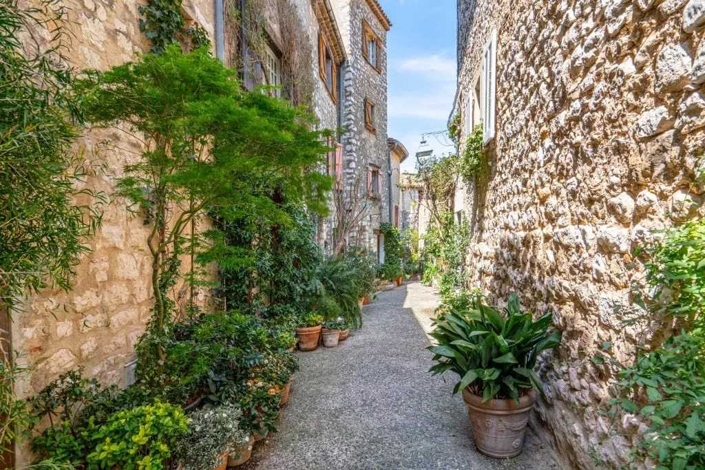 Photo of a cobblestone street in Saint-Paul-de-Vence France, with stone buildings on either side and green plants along the street--don't miss this stop during your south of France itinerary!