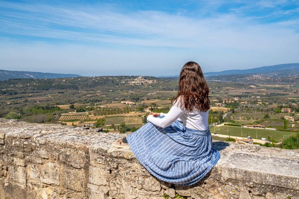 Kate in a blue skirt sitting on a stone wall overlooking the countryside of the Luberon Valley. Taken in Bonnieux France.