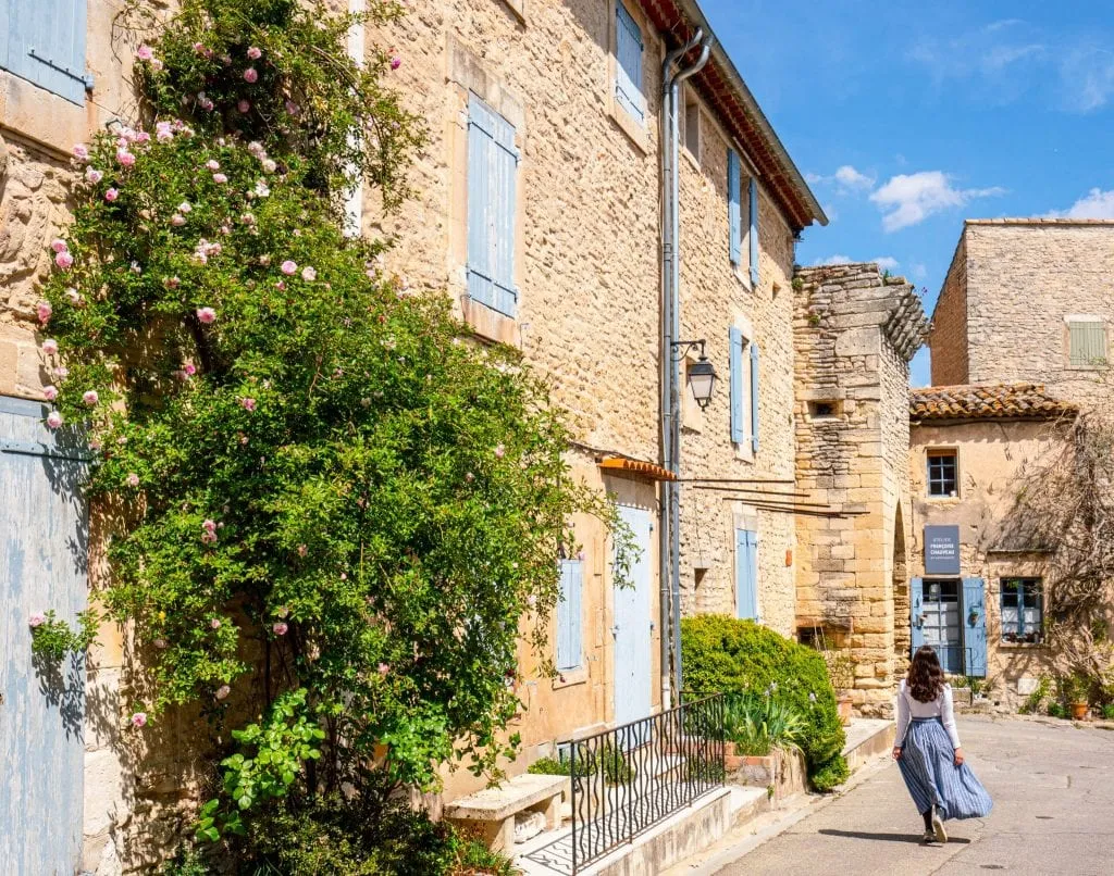 Kate in a blue skirt in Goult, France. There's a stone building with blue shutters to her left, and she's facing away from the camera. Goult is one of the best places to visit in the south of France.