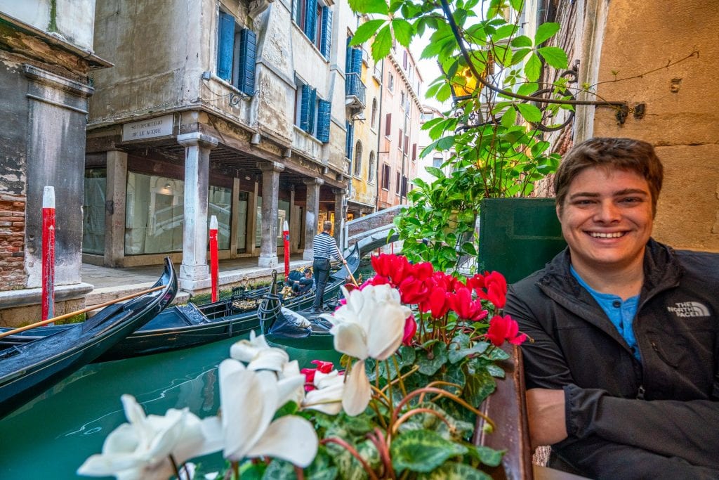 Man sitting in a restaurant along a Venetian canal. There's a flowerbox in the window next to him.