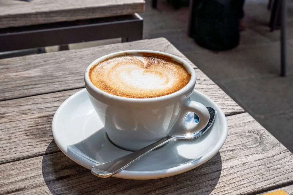 Cappuccino on a plate with a spoon next to it on a wooden table bathed in sunlight. Excellent Italy travel tip: start every morning with one of these!