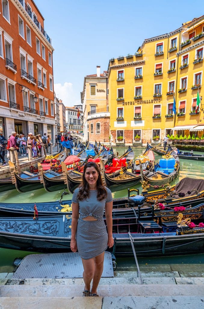 Kate Storm in a striped dress standing in front of a crowd of gondolas near Piazza San Marco with a yellow buidling behind her--if you want to have peaceful gondola ride in Venice, this isn't the best place to get on!