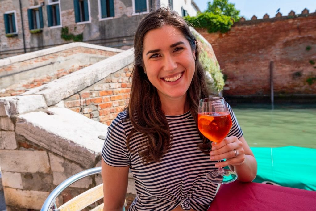 Kate Storm in Venice wearing a striped dress and holding up an Aperol Spritz