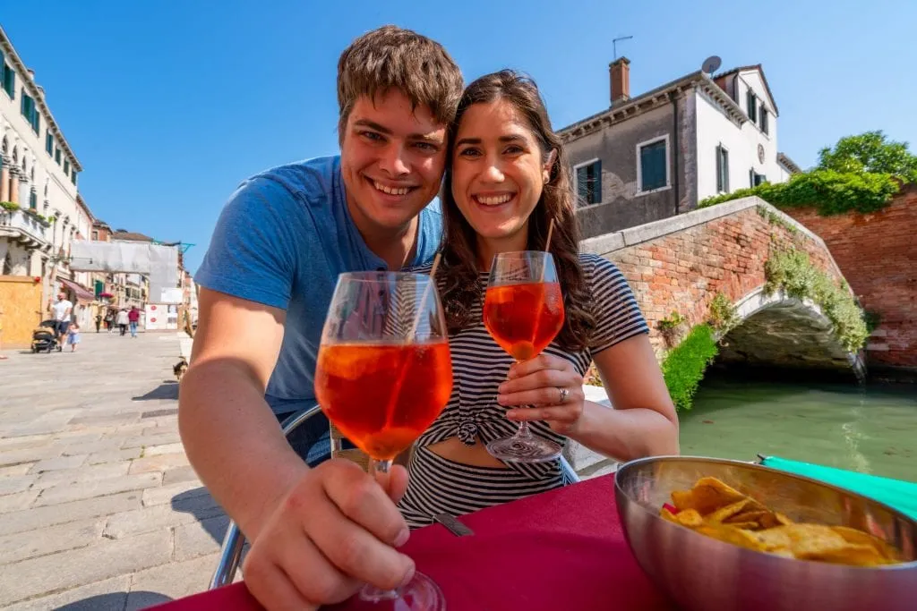 Couple sitting at a table in Cannaregio, Venice, holding aperol spritzes.