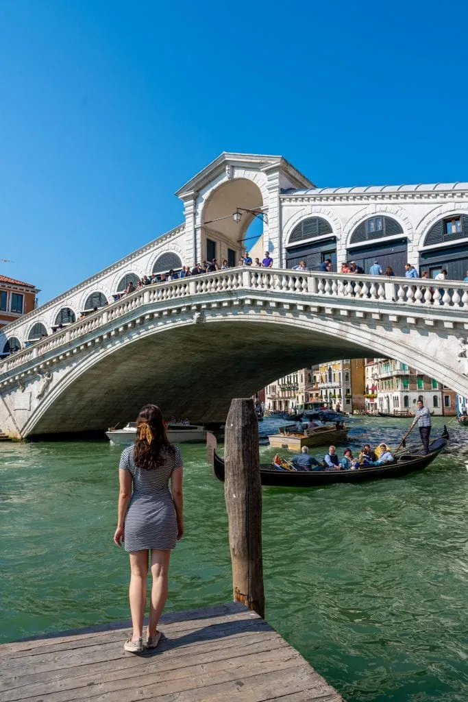 Kate Storm standing on a dock looking up at the Rialto Bridge in Venice. One of the coolest Venice history facts is that this bridge is the oldest across the grand canal