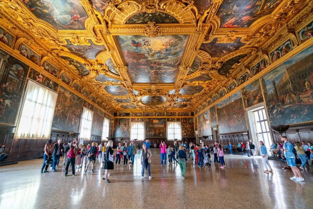 Photo of interior of largest council chamber in the Doge's Palace--definitely worth visiting during one day in Venice! The ceiling is the focus of the shot and is covered in gold, there's a crowd in the distance.