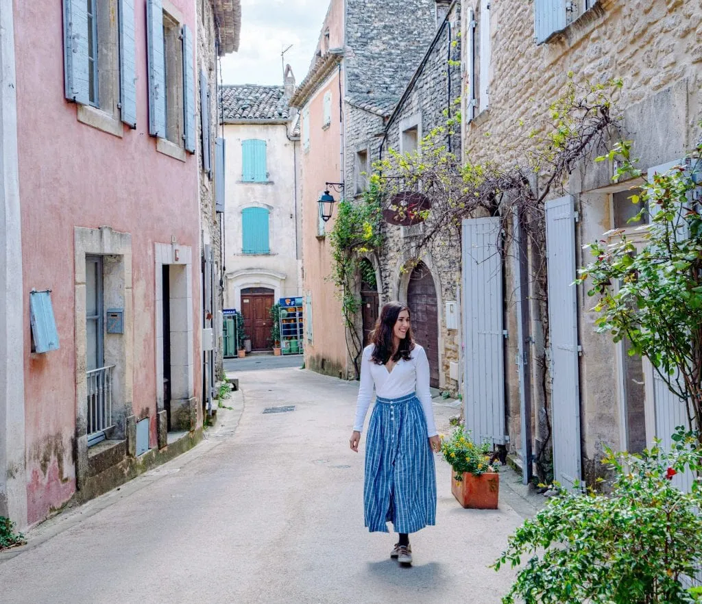 Kate Storm walking down a narrow street in Goult during our France road trip. There's a pink building to her left and she's wearing a long blue skirt.