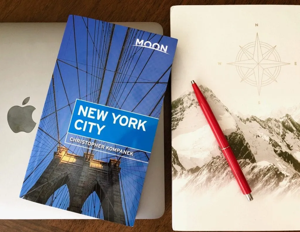 Photo of a Macbook Pro, a notebook with mountains on the cover, and a red pen. A copy of Moon New York City is laying on top of them--use this to find some of the best things to do in MIdtown NYC!