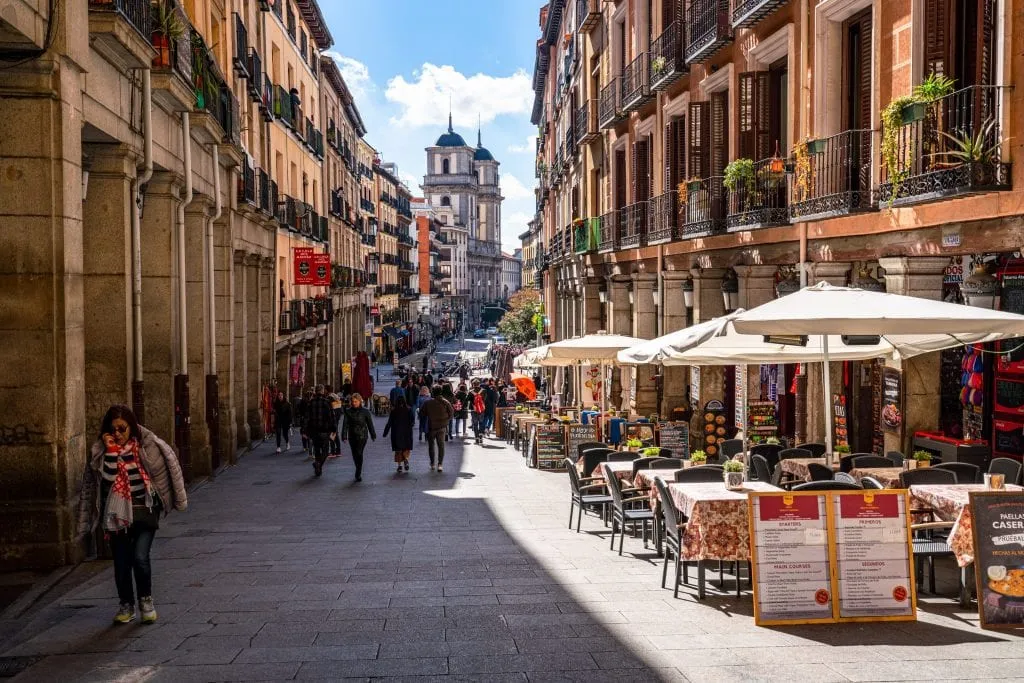 Photo of a quiet street in Madrid Spain, with tables that have umbrellas set over them on the righthand side.