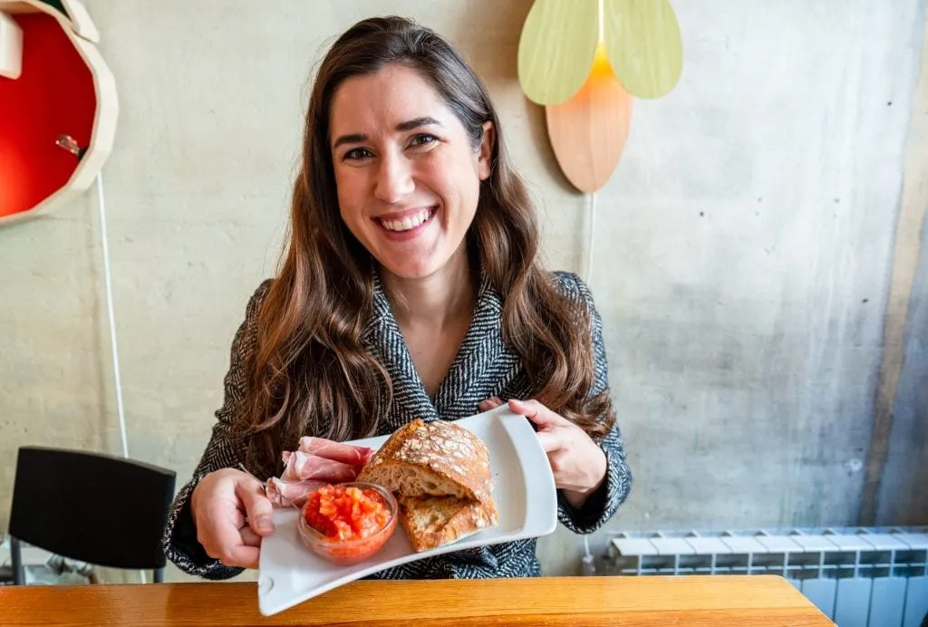 Kate Storm in a gray coat holding up a plate of bread, tomato, and ham--el b_us is a great stop for breakfast during your 3 days in Madrid itinerary!