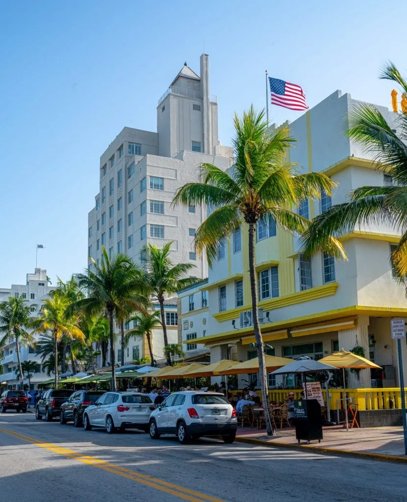 Ocean Drive in Miami Beach, a must see during any trip to Miami! The street is lined with palm trees and there's a yellow and white building in the foreground. An American flag is waving at the top of the photo.