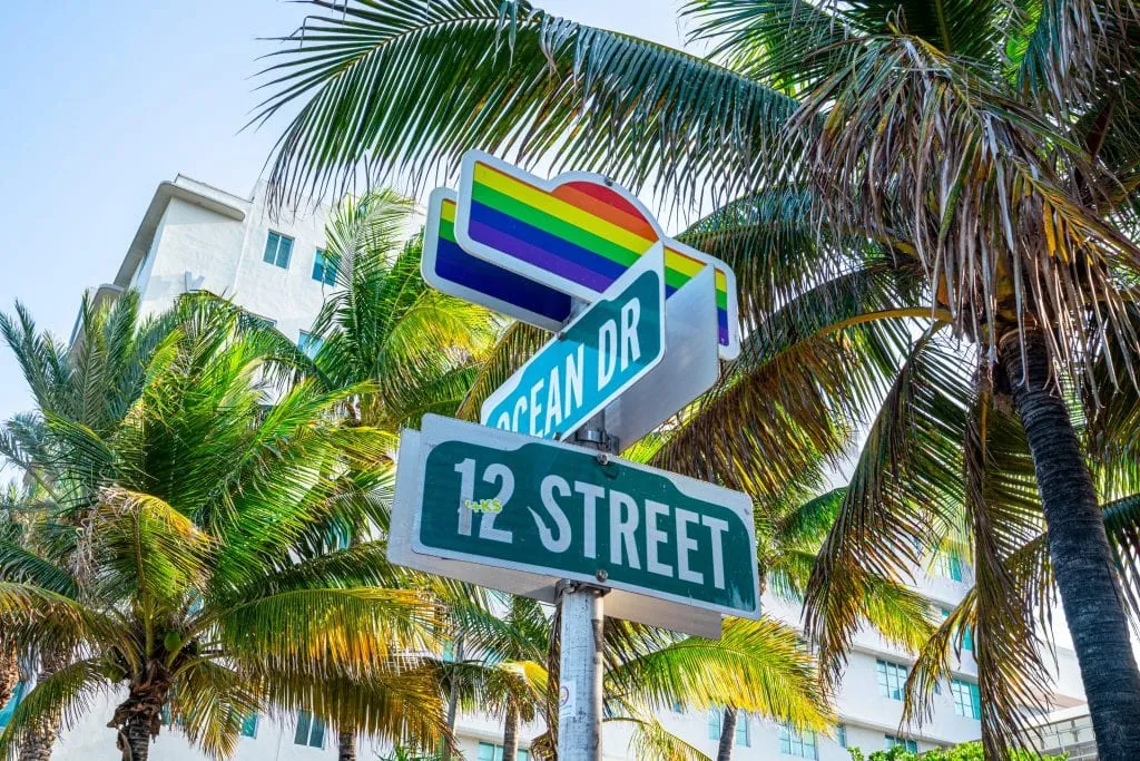 Photo of the sign for cross streets Ocean Drive and 12th Street. There's pride rainbow on top of the sign and palm trees in the background.