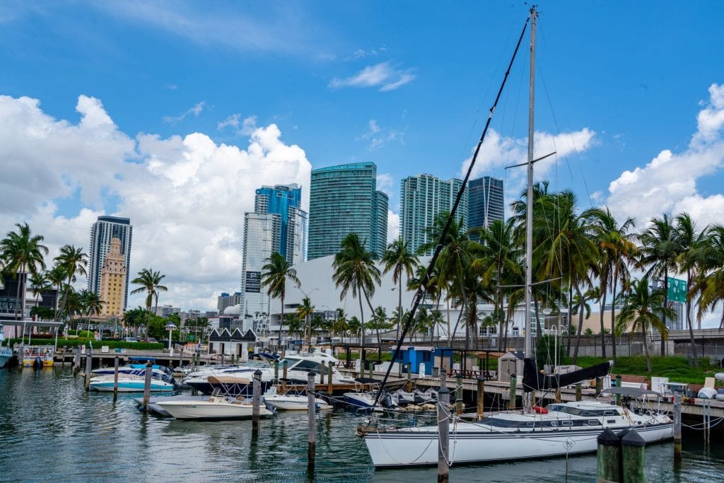 Harbor in Miami. There's a sailboat in the foreground, smaller boats behind it, and skyscrapers visible beyond that.