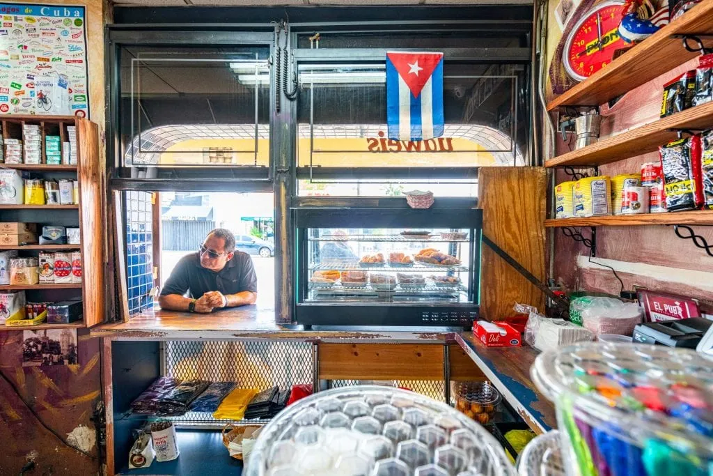 Man standing at the window of El Pub in Little Havana waiting for coffee. There's a case of empanadas to the right and a Cuban flag hanging near the top right of the photo.