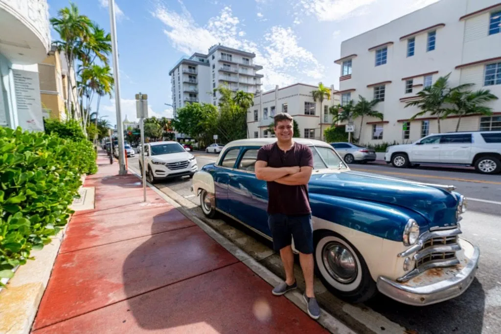 Jeremy standing next to a classic car parked on Collins Ave. There's a sidewalk to the left that is painted maroon.