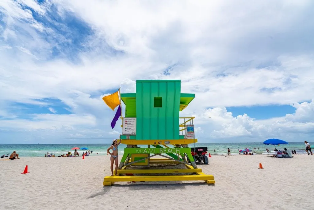 Kate standing on a green art deco lifeguard stand on South Beach--an essential place to visit during any 3 day Miami itinerary! The lifeguard stand says "Miami Beach" and has two flags on it.