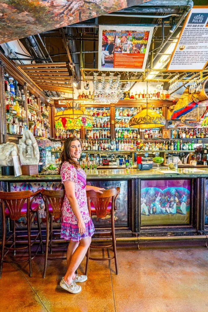 Kate in a pink dress standing in Cubaocho, in front of a historic wooden bar with many glass bottles and hanging lamps. Don't skip this during your long weekend in Miami!